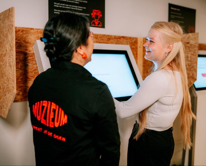 Een lachende bezoeker samen met een medewerker staat bij het grote digitale scherm waarop een experiment gedaan kan worden.