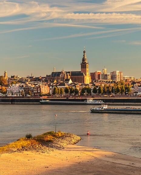 Overzicht op de Stevenskerk en de stad Nijmegen vanaf de andere kant van de waal. Op de voorgrond zie je zand, de waal en twee boten.