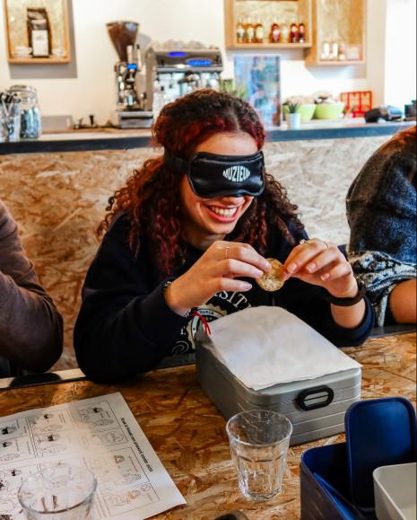 Een bezoeker met blinddoek op, die een koekje in de handen heeft en eraan voelt.